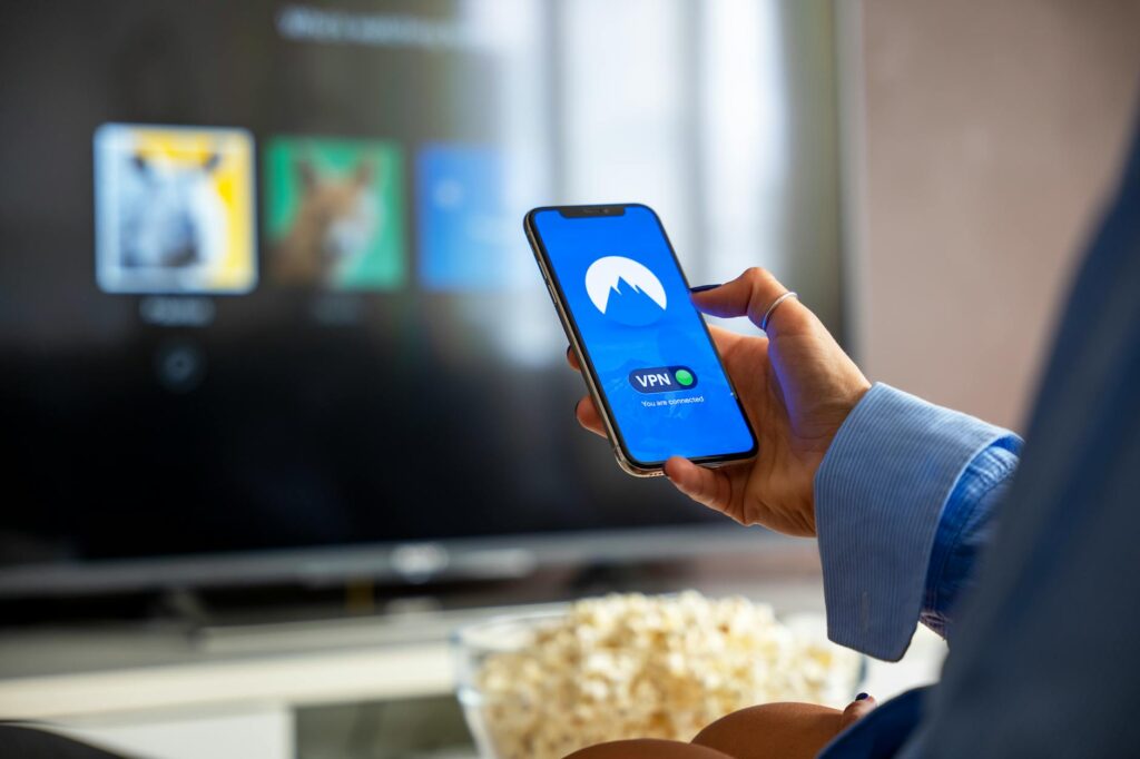 Person using VPN on smartphone while watching smart TV at home. (Photo by Stefan Coders on Pexels)