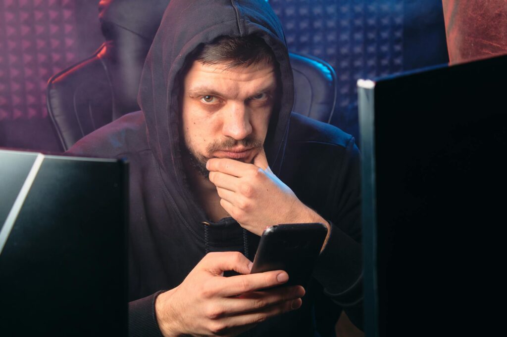 A man in a black hoodie contemplating while using a smartphone, surrounded by digital screens. (Photo by Mikhail Nilov on Pexels)